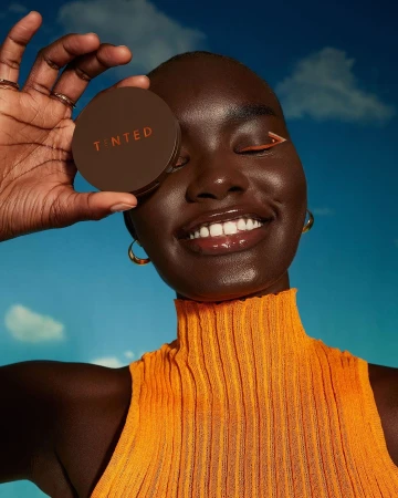 Smiling woman with dark skin holds brown tinted foundation compact. She wears an orange top and gold hoops against a cloudy sky.