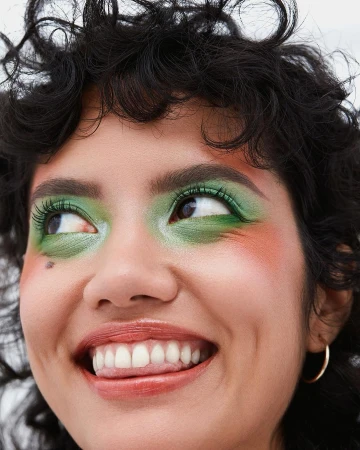 Close-up portrait of a woman with green eyeshadow and dark curly hair looking upward with a slight smile