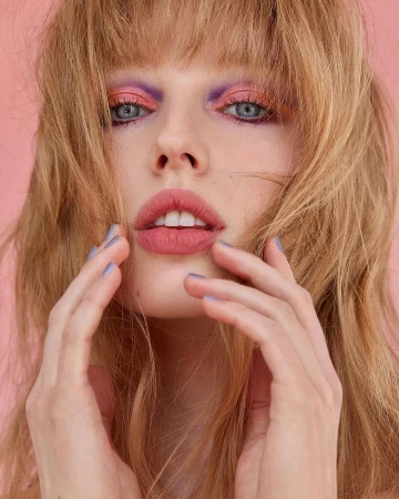 Close-up of a woman with light skin and strawberry blonde hair. Her makeup is lavender and pink. She has her hands raised to her face.