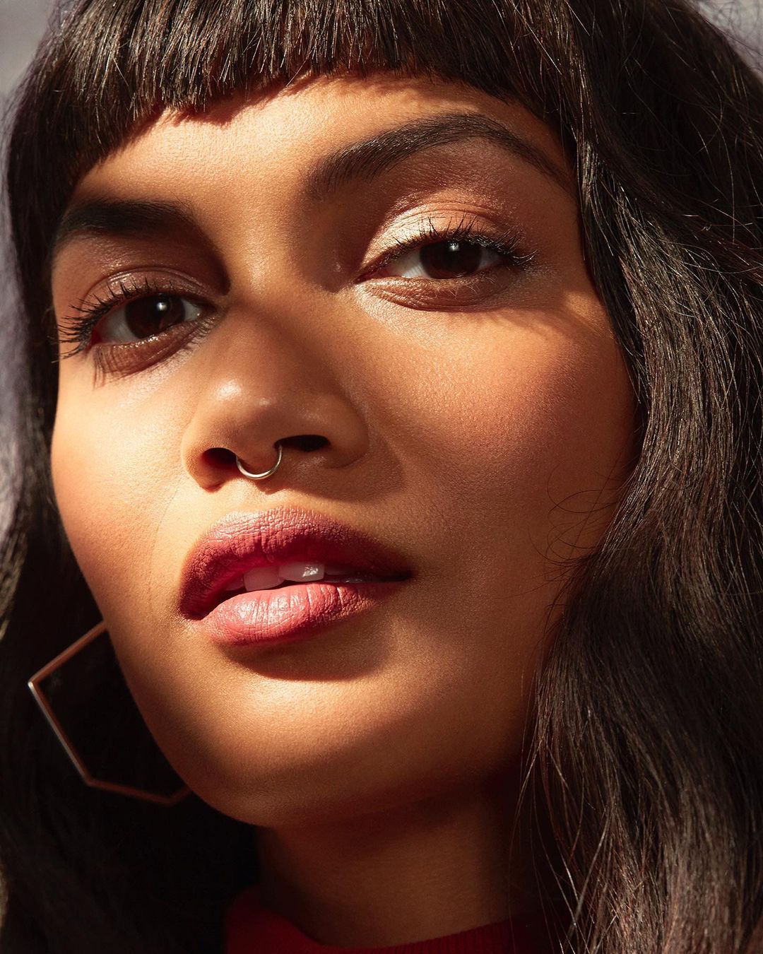Close-up portrait of an adult female with dark hair and a septum piercing, illuminated by natural light casting shadows on her face.