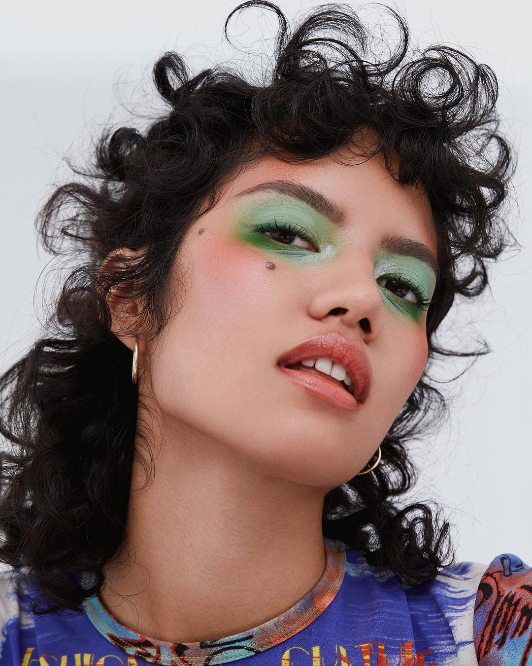 Close-up portrait of a woman with creative green eyeshadow and curly dark hair against a white backdrop. Beauty shot, makeup.