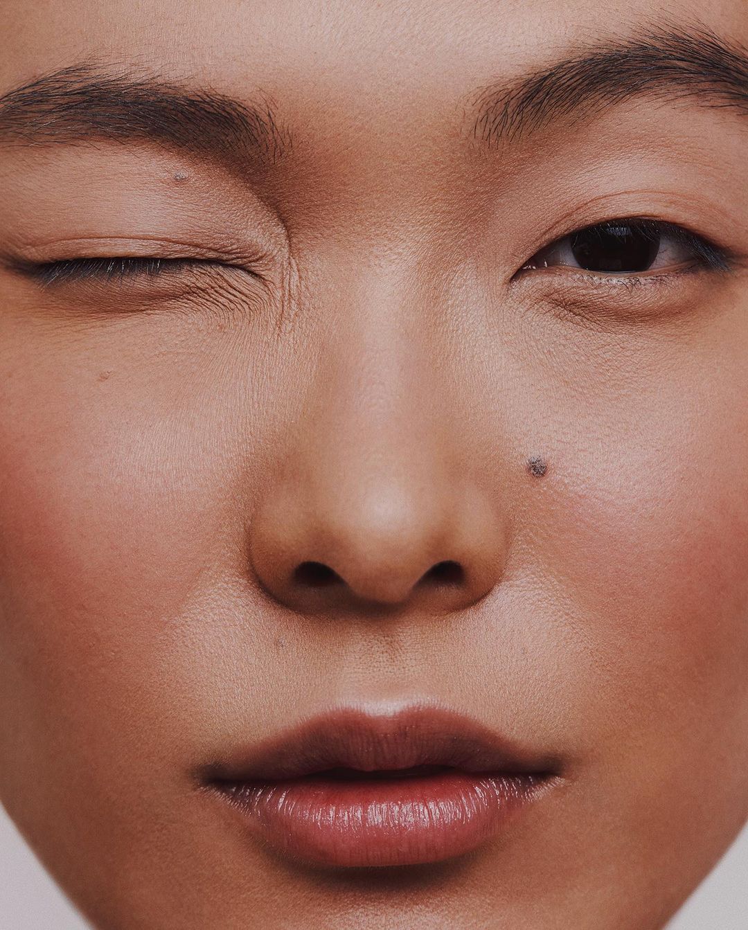 Extreme close-up portrait of an Asian woman winking, showcasing detailed skin texture, moles, and natural makeup.