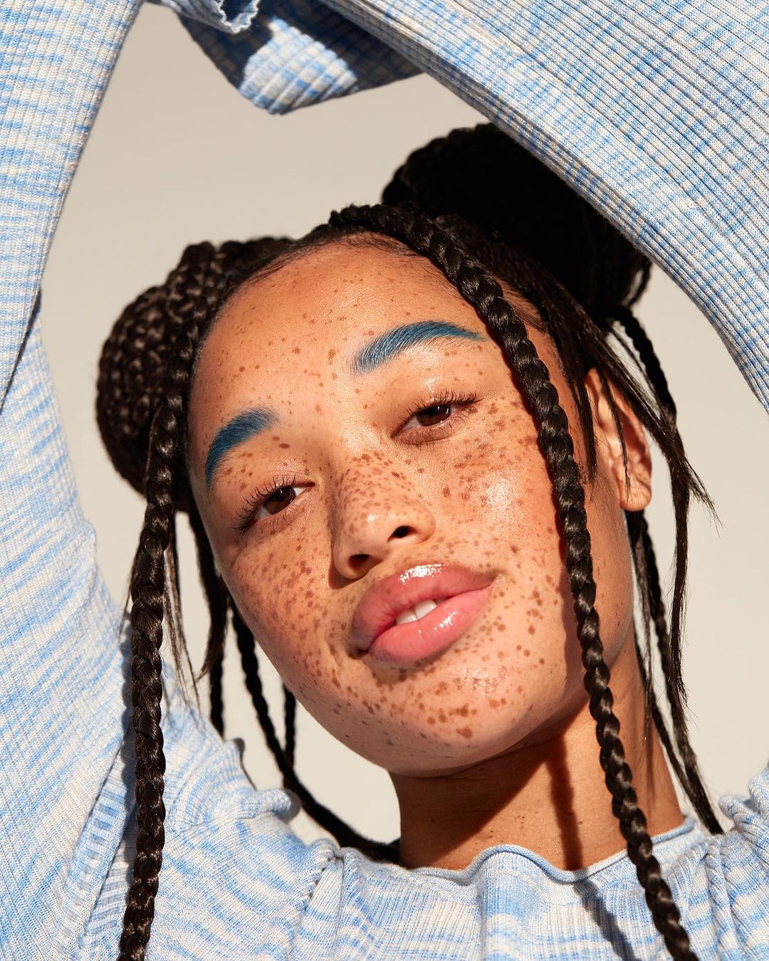 Close-up portrait of a young adult female model with freckles and blue eyebrows, wearing a textured blue and white top in a studio setting.