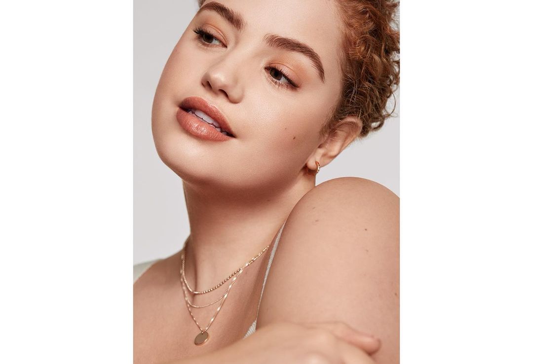 Close-up shot of an adult woman with curly auburn hair, fair skin, wearing layered gold necklaces against a plain white backdrop.
