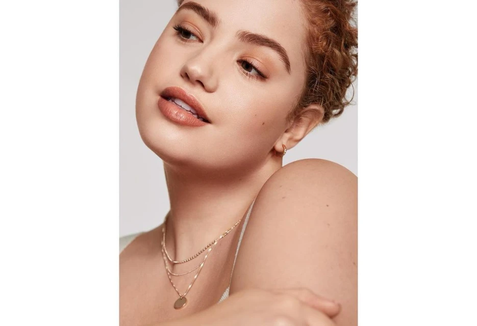 Close-up shot of an adult woman with curly auburn hair, fair skin, wearing layered gold necklaces against a plain white backdrop.