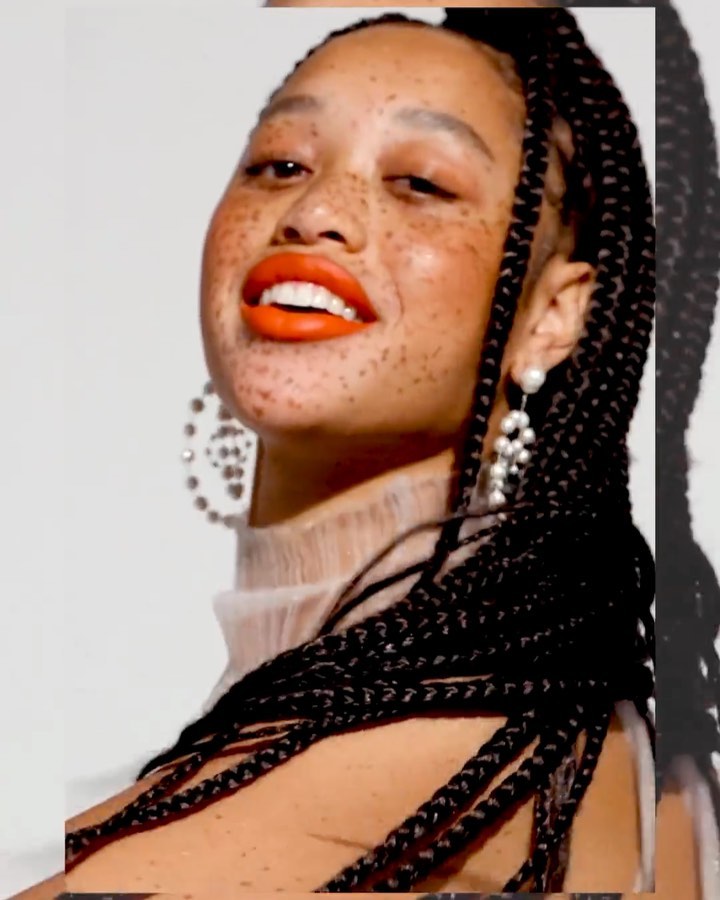 Close-up studio shot of a fashionable young adult female with freckles, wearing a white top, orange lipstick, and pearl earrings, smiling.