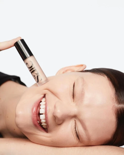 A smiling woman with a makeup product. Beauty shot of a woman with light skin, eyes closed, and fair skin, holding a SAIE product.