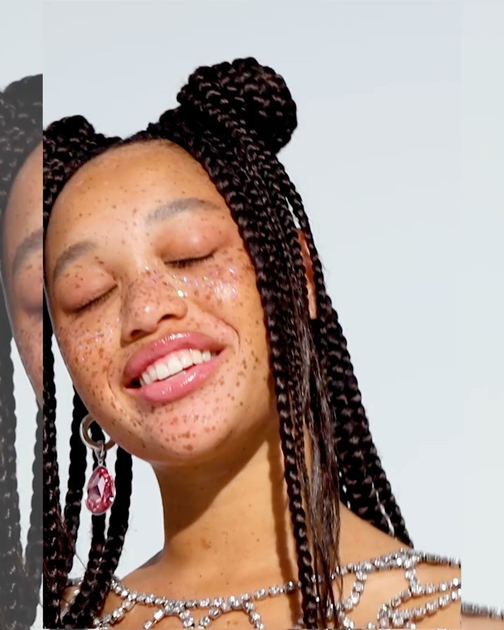 Close-up of smiling woman with glitter freckles, braided hair buns, statement earrings, and a jeweled top against a white background.
