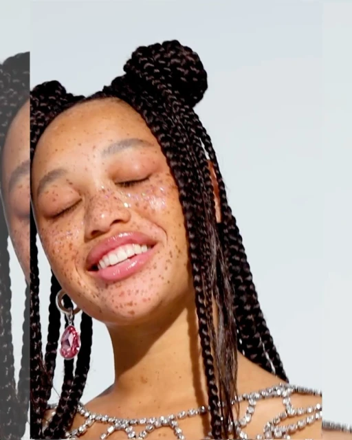 Close-up of smiling woman with glitter freckles, braided hair buns, statement earrings, and a jeweled top against a white background.
