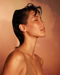 Profile portrait of a woman with wet-look dark hair and freckles against a warm sunset sky background