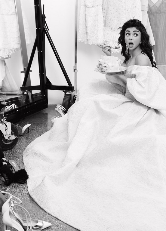 Sarah Hyland in off-shoulder gown eating cake on set surrounded by shoes and dresses, monochromatic, high-angle view.