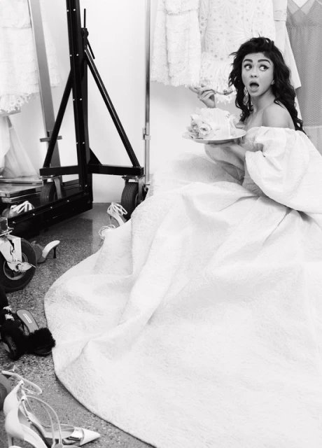 Sarah Hyland in off-shoulder gown eating cake on set surrounded by shoes and dresses, monochromatic, high-angle view.