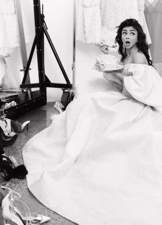 Sarah Hyland in off-shoulder gown eating cake on set surrounded by shoes and dresses, monochromatic, high-angle view.