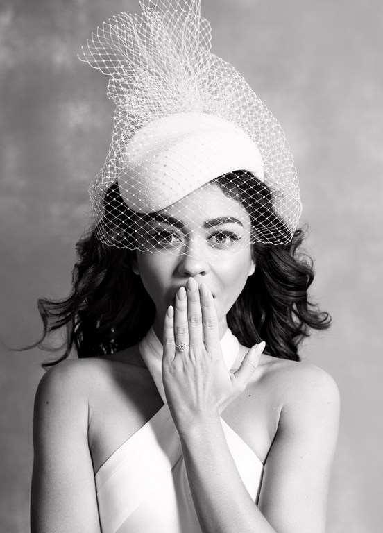 Woman in a white dress and hat with netting poses with her hand near her mouth against a neutral background in a medium shot.