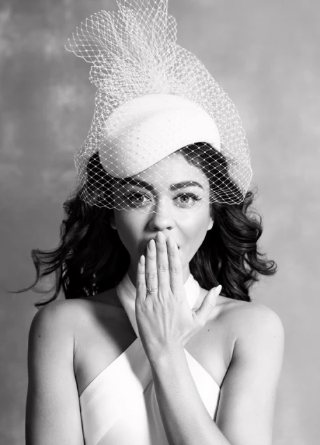 Woman in a white dress and hat with netting poses with her hand near her mouth against a neutral background in a medium shot.