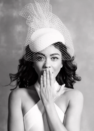 Woman in a white dress and hat with netting poses with her hand near her mouth against a neutral background in a medium shot.