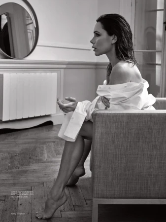 Victoria Beckham seated in an armchair wearing an off-the-shoulder white cotton shirt in a room with a round mirror