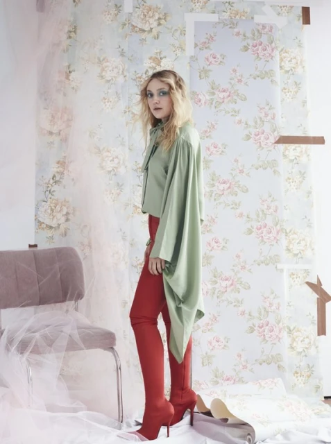Adult female fashion model in an indoor photo studio wearing red thigh-high boots, a pastel green top, posing in front of floral wallpaper.