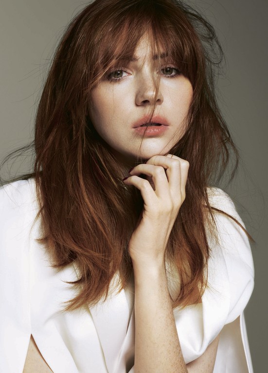 Portrait of a woman with red hair and bangs wearing a white top with her hand near her chin, against a solid grey background.