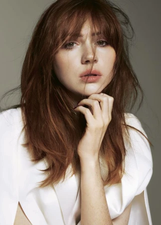 Portrait of a woman with red hair and bangs wearing a white top with her hand near her chin, against a solid grey background.