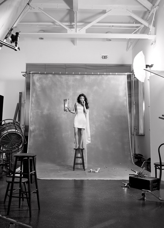 A young woman stands on a stool in a photo studio, wearing a dress and eating from a snack bag against a gray backdrop.