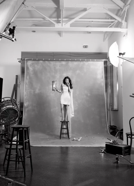 A young woman stands on a stool in a photo studio, wearing a dress and eating from a snack bag against a gray backdrop.