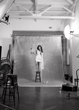 A young woman stands on a stool in a photo studio, wearing a dress and eating from a snack bag against a gray backdrop.