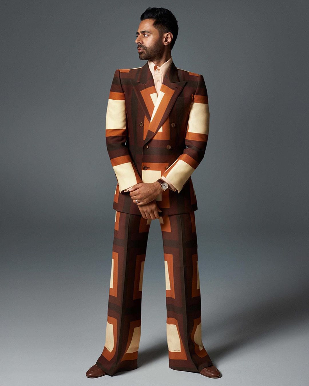 Full-length portrait of adult male with dark hair and beard, wearing a retro-patterned brown suit, posing against a grey backdrop.