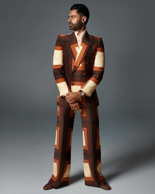 Full-length portrait of adult male with dark hair and beard, wearing a retro-patterned brown suit, posing against a grey backdrop.
