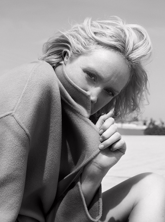 Close-up black and white portrait of a blonde woman pulling up a heavy collar of her coat to partially cover her face.