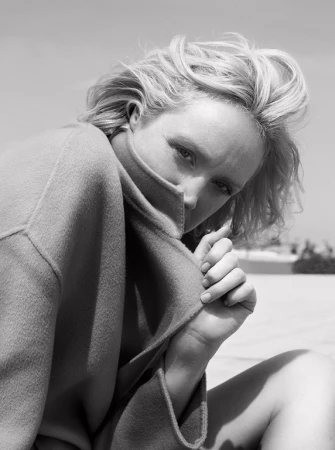 Close-up black and white portrait of a blonde woman pulling up a heavy collar of her coat to partially cover her face.