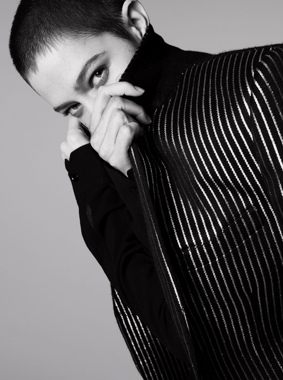 Monochrome portrait of a young adult woman with hands near her face, wearing a striped top and a black turtleneck against a grey background.