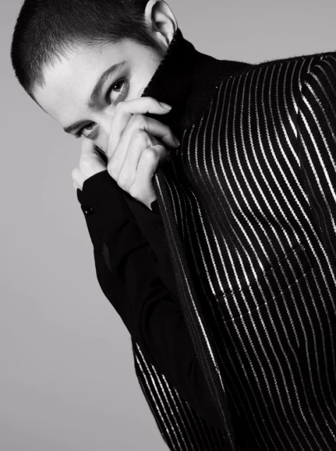 Monochrome portrait of a young adult woman with hands near her face, wearing a striped top and a black turtleneck against a grey background.