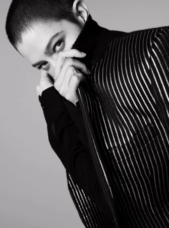 Close-up portrait of a model with short cropped hair pulling up a striped jacket collar to partially cover her face.