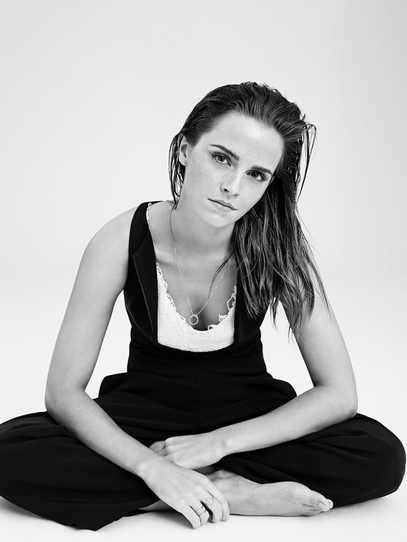 Black and white studio portrait of a young adult woman with wet hair sitting cross-legged, wearing a black jumpsuit with a white lace top.