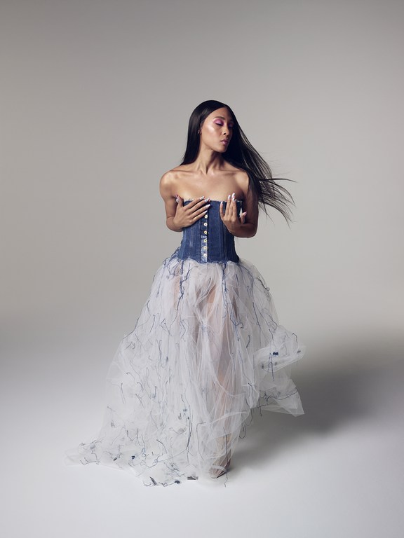 Woman in a denim and tulle dress poses in a photo studio with a neutral backdrop. The model has long black hair and fair skin.