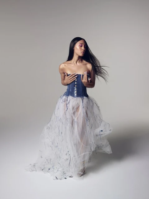 Woman in a denim and tulle dress poses in a photo studio with a neutral backdrop. The model has long black hair and fair skin.
