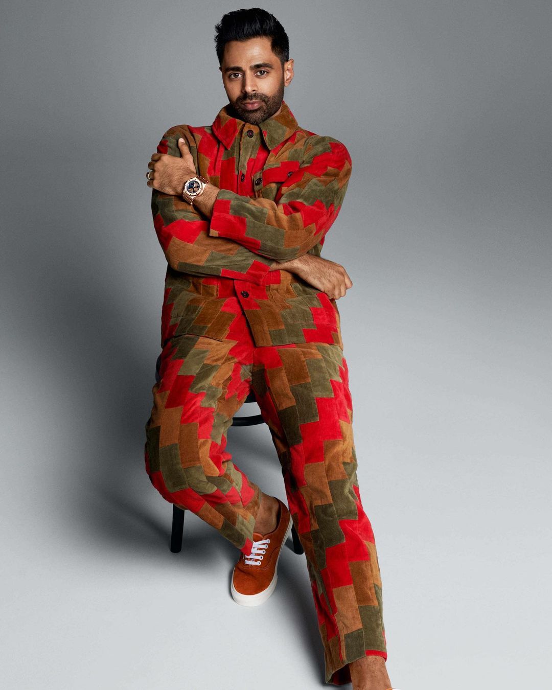 Hasan Minhaj poses in a colourful patchwork suit, seated indoors, medium shot, looking at camera, studio setting on neutral grey background.