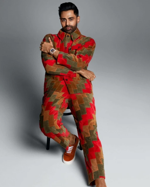 Hasan Minhaj poses in a colourful patchwork suit, seated indoors, medium shot, looking at camera, studio setting on neutral grey background.