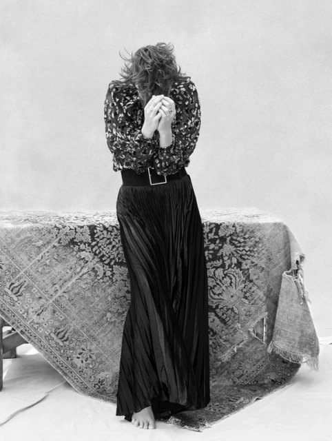 Monochrome portrait of a woman covering her face with hands, wearing a patterned top and long black skirt, standing behind a vintage table.