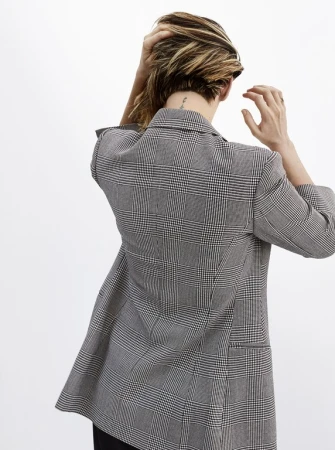 Woman in grey plaid blazer, facing away from camera, arranges her highlighted hair against a plain white background in a studio setting.