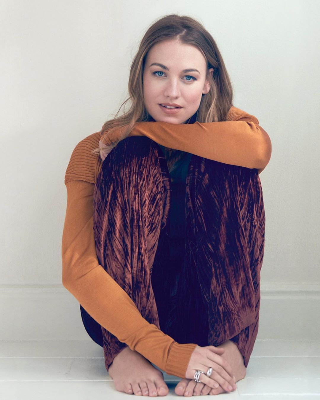 Attractive woman with long light-brown hair, wearing a mustard-colored sweater and crushed-velvet pants, sits indoors against a white wall.