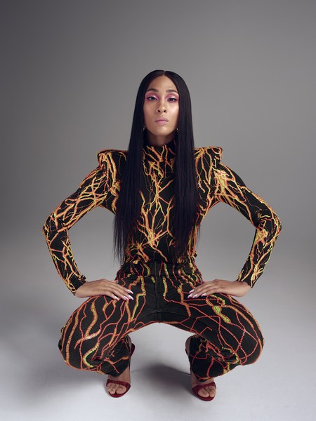 Adult woman squats in a long-sleeved jumpsuit with orange lightning bolt pattern against a grey studio background with red high heels.
