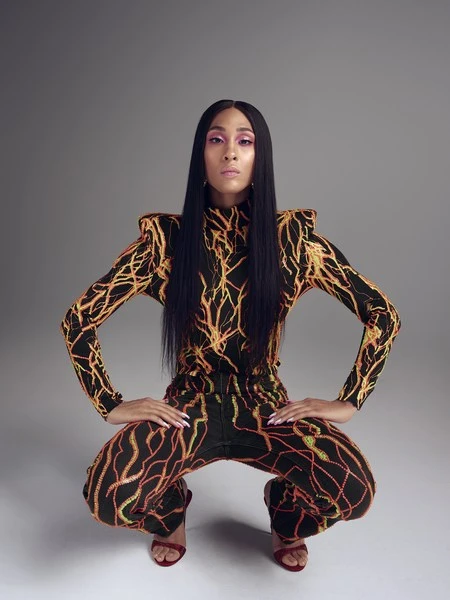 Adult woman squats in a long-sleeved jumpsuit with orange lightning bolt pattern against a grey studio background with red high heels.