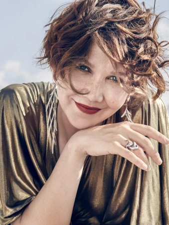 Close-up portrait of Maggie Gyllenhaal with short curly hair, smiling and resting her chin on her hand, wearing a metallic gold top.