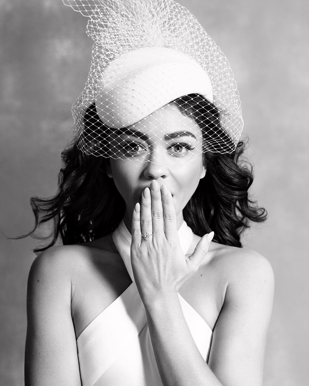Black and white portrait of a female wearing a white dress, hat, and netting, with her hand covering her mouth and her ring visible.