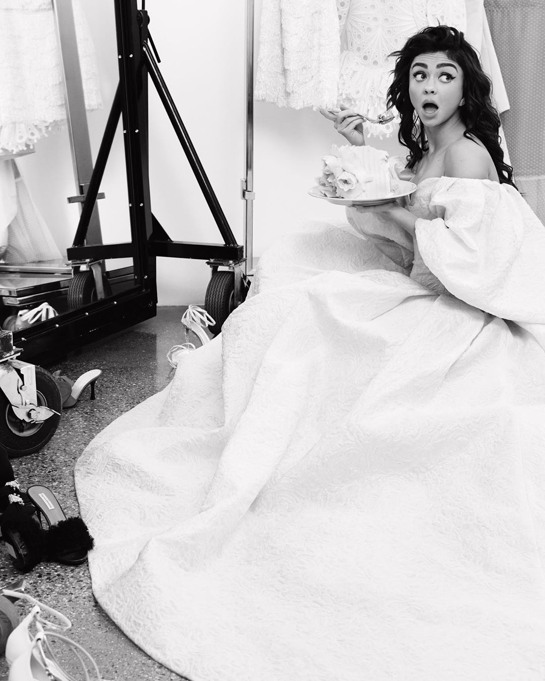 Black and white portrait of a young adult woman in a gown holding a cake, inside what looks like a photo studio.