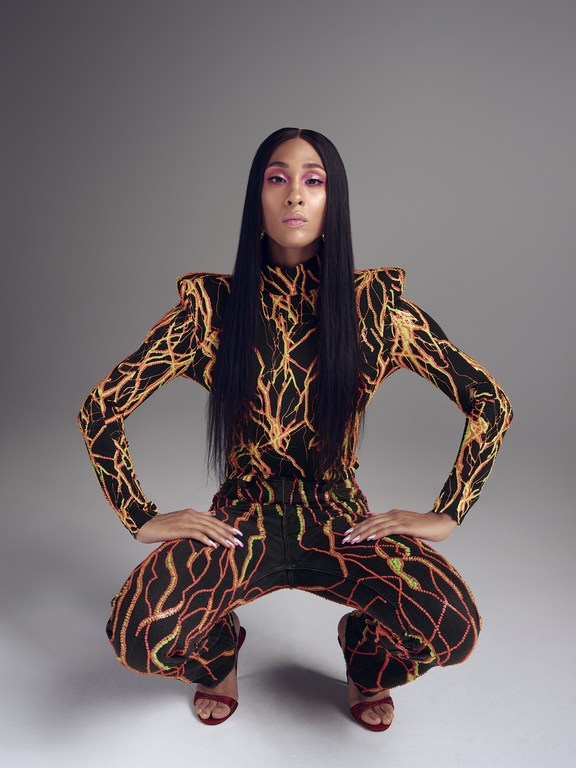 Woman with long black hair poses crouching in patterned, long-sleeved jumpsuit and red heels against a neutral grey studio backdrop.