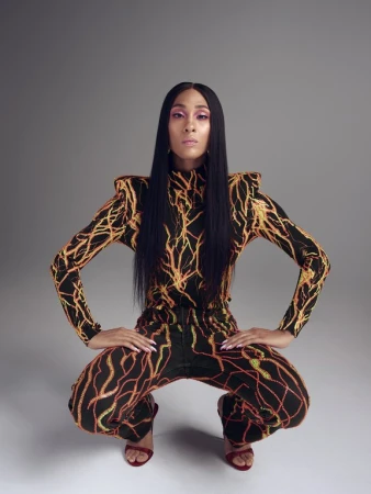 Woman with long black hair poses crouching in patterned, long-sleeved jumpsuit and red heels against a neutral grey studio backdrop.