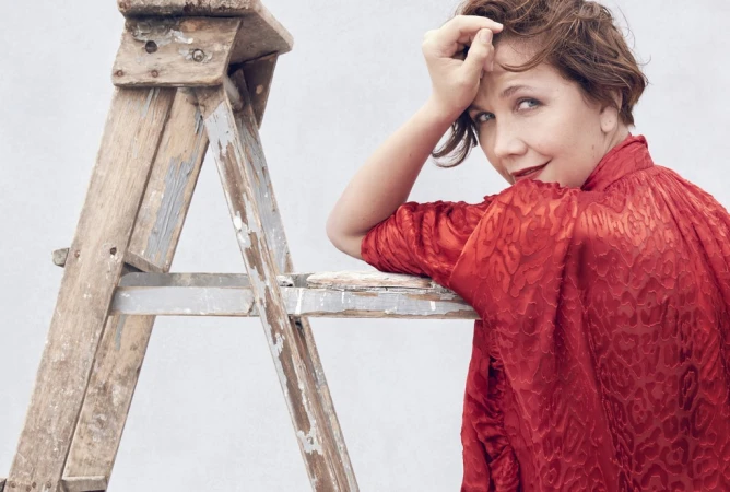 Woman with short brown hair wearing a patterned red blouse leaning against a wooden step ladder against a light grey wall.